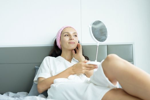 A beautiful woman wearing a towel and a white bathrobe and pink headband has to look a mirror with happy and relaxing on the bed at a condominium in the morning.