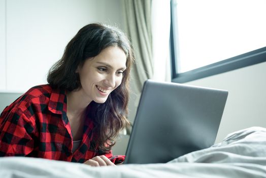 Beautiful woman working on a laptop with smiling and lying down on the bed at a condominium in the morning.