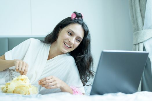 A beautiful woman eating snack potato and playing laptop with lying down on the bed and happiness at a condominium in the morning.