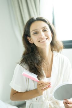 A beautiful woman wearing a towel and a white bathrobe has to look a mirror for combing hair with a pink comb and on the bed at a condominium in the morning.