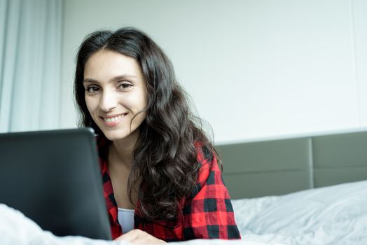 Beautiful woman working on a laptop with smiling and lying down on the bed at a condominium in the morning.