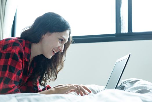 Beautiful woman working on a laptop with smiling and lying down on the bed at a condominium in the morning.