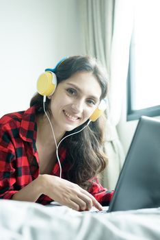 Beautiful woman working and listening to music on a laptop with yellow headphone and lying down on the bed at a condominium in the morning.