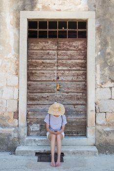 Beautiful young female tourist woman sitting and resting on vinatage wooden doorstep and textured stone wall at old Mediterranean town, smiling, holding, using smart phone to network on vacationes.