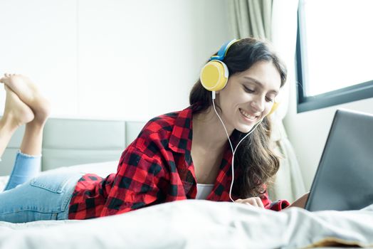 Beautiful woman working and listening to music on a laptop with yellow headphone and lying down on the bed at a condominium in the morning.