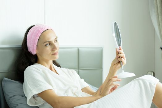 A beautiful woman wearing a towel and a white bathrobe and pink headband has to look a mirror with happy and relaxing on the bed at a condominium in the morning.