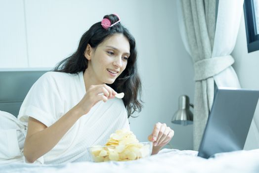 A beautiful woman eating snack potato and playing laptop with lying down on the bed and happiness at a condominium in the morning.