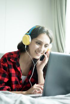 Beautiful woman working and listening to music on a laptop with yellow headphone and lying down on the bed at a condominium in the morning.