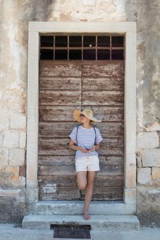 Beautiful young female tourist woman standing in front of vinatage wooden door and textured stone wall at old Mediterranean town, smiling, holding, smart phone to network on vacationes.