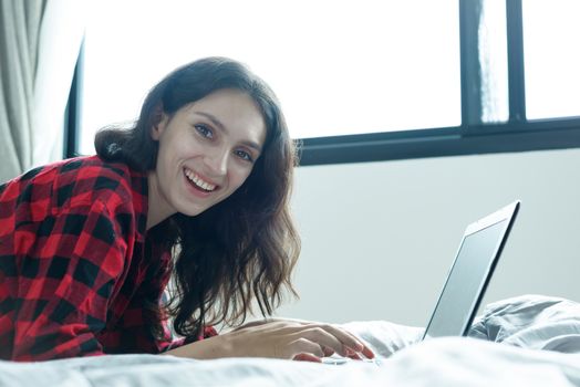 Beautiful woman working on a laptop with smiling and lying down on the bed at a condominium in the morning.