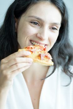 A beautiful woman wearing a towel and a white bathrobe has to eat a pizza with happy and relaxing on the bed at a condominium in the morning.