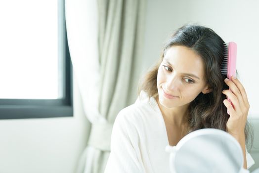 A beautiful woman wearing a towel and a white bathrobe has to look a mirror for combing hair with a pink comb and on the bed at a condominium in the morning.