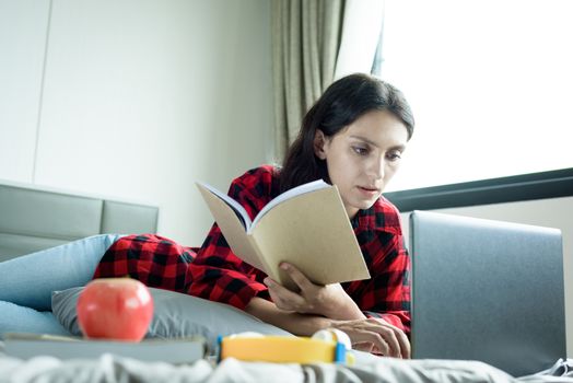 Beautiful woman working and reading a book on a laptop and lying down on the bed at a condominium in the morning.