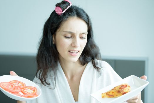 A beautiful woman wearing a towel and a white bathrobe has to eat a pizza and sliced tomato with happy and relaxing on the bed at a condominium in the morning.