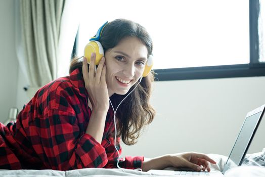 Beautiful woman working and listening to music on a laptop with yellow headphone and lying down on the bed at a condominium in the morning.