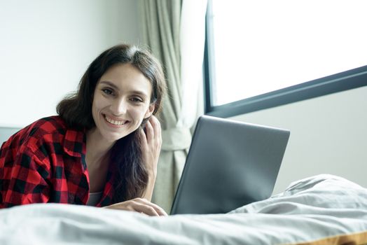 Beautiful woman working on a laptop with smiling and lying down on the bed at a condominium in the morning.