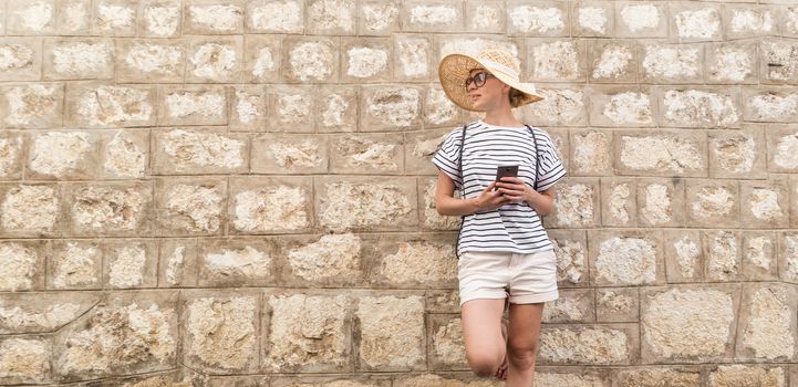 Beautiful young female tourist woman standing in front of old textured stone wall at old Mediterranean town, smiling, holding, smart phone to network on vacationes. Copy space.