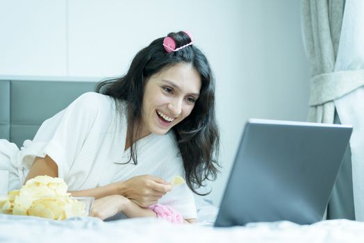 A beautiful woman eating snack potato and playing laptop with lying down on the bed and happiness at a condominium in the morning.