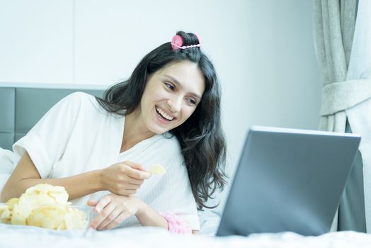 A beautiful woman eating snack potato and playing laptop with lying down on the bed and happiness at a condominium in the morning.