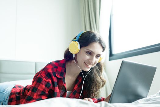 Beautiful woman working and listening to music on a laptop with yellow headphone and lying down on the bed at a condominium in the morning.