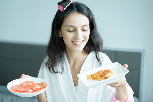 A beautiful woman wearing a towel and a white bathrobe has to eat a pizza and sliced tomato with happy and relaxing on the bed at a condominium in the morning.