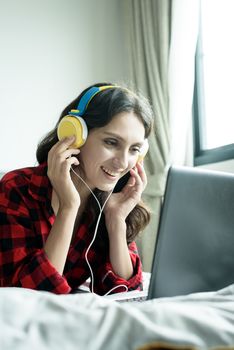 Beautiful woman working and listening to music on a laptop with yellow headphone and lying down on the bed at a condominium in the morning.