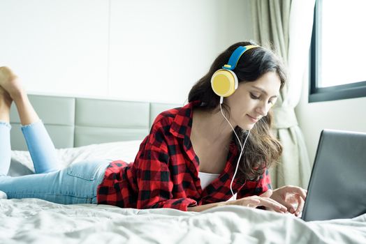 Beautiful woman working and listening to music on a laptop with yellow headphone and lying down on the bed at a condominium in the morning.