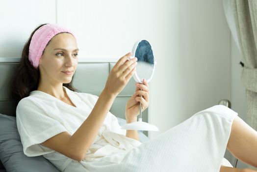 A beautiful woman wearing a towel and a white bathrobe and pink headband has to look a mirror with happy and relaxing on the bed at a condominium in the morning.