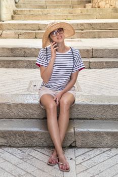 Beautiful young tourist woman at summer vacations, sitting on old stone steps of old medieval Mediterranean costal town, smiling, talking on phone with her loved ones at home.