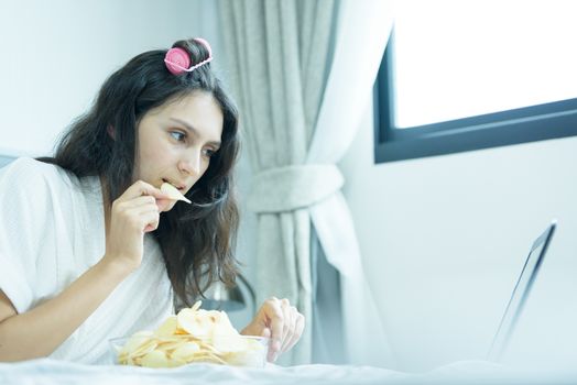 A beautiful woman eating snack potato and playing laptop with lying down on the bed and happiness at a condominium in the morning.