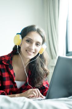 Beautiful woman working and listening to music on a laptop with yellow headphone and lying down on the bed at a condominium in the morning.