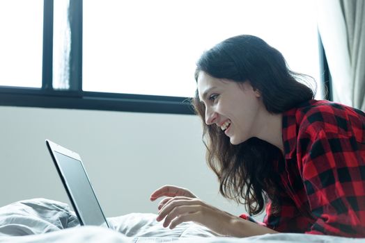 Beautiful woman working on a laptop with smiling and lying down on the bed at a condominium in the morning.