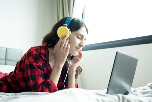 Beautiful woman working and listening to music on a laptop with yellow headphone and lying down on the bed at a condominium in the morning.