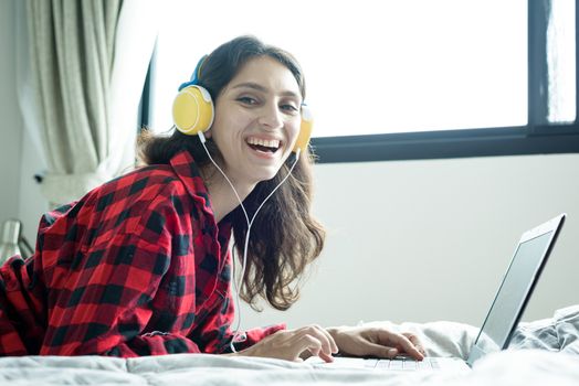 Beautiful woman working and listening to music on a laptop with yellow headphone and lying down on the bed at a condominium in the morning.