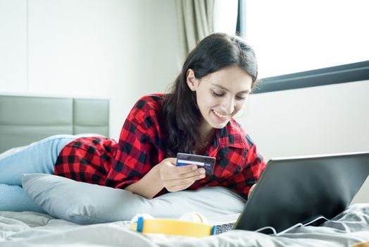 Beautiful woman shopping online on a laptop with the credit card and lying down on the bed at a condominium in the morning.