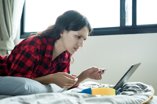 A beautiful woman has lain down on the bed at a condominium and shopping online on a laptop by the credit card with unhappy and worried in the morning.