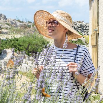 Beautiful blonde young female traveler wearing straw sun hat enjoying summer on Mediterranean cost strolling among lavender flowers on traditional costal village garden.