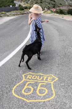 Young attractive female traveler wearing striped summer dress and straw hat standing on an endless straight empty road in the middle of nowhere on the Route 66 road and feeding black sheep.