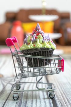 Beautifully iced cupcake with purple frosting and black chocolate base on a shopping cart on a wooden floor with blurred out background. Shows hobbies and skills people are leanring in the coronavirus pandemic