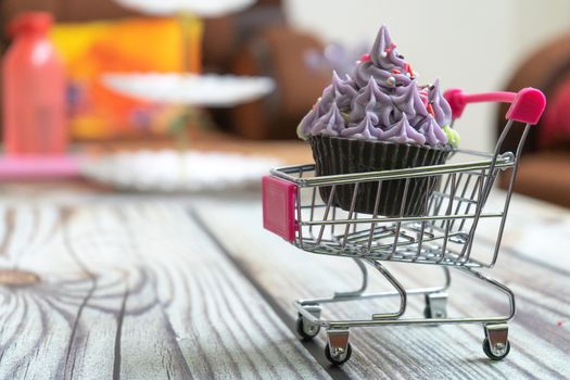 Beautifully iced cupcake with purple frosting and black chocolate base on a shopping cart on a wooden floor with blurred out background. Shows hobbies and skills people are leanring in the coronavirus pandemic