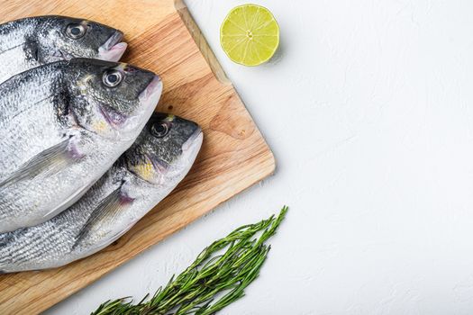 Raw dorada or sea bream fish set on chopping board with herbs for grill uncooked on white textured background top view .