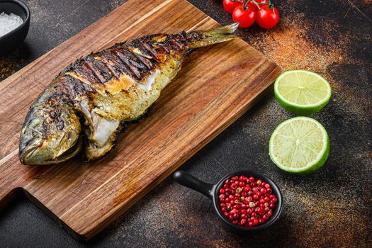 Grilled sea bream or dorado raw fish on chopping board with ingredients over old metall dark background, side view with space for text