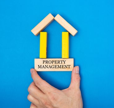 Male hand placing a property management in a house made of wooden blocks in a conceptual image of real estate market. Over blue background.