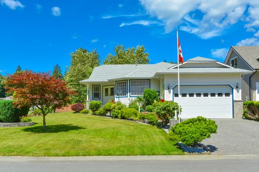 One level house with Canadian flag on a front yard on blue sky background. Big house with lawn in front and mountain view background. British Columbia, Canada.