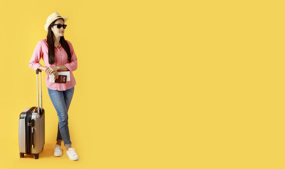 Asian women long hair wear Straw hat with black ribbon, sunglasses in hand holding passport book and travel bag with copy space banner. Young girl ready to travel with suitcase and passport on yellow background. Summer travel concept.