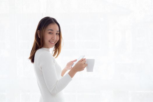 Beautiful young woman in white sweater drinking coffee and holding smartphone at a window.