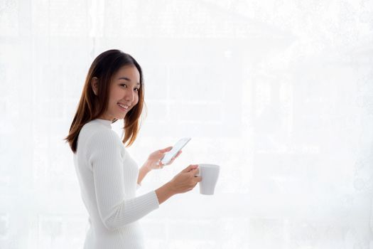 Beautiful young woman in white sweater drinking coffee and holding smartphone at a window.