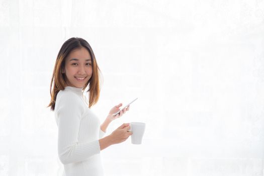 Beautiful young woman in white sweater drinking coffee and holding smartphone at a window.