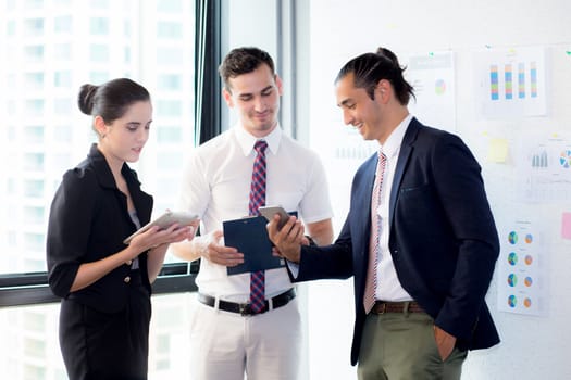 Three businesspeople standing in modern office looking at smart mobile phone and talking in meeting room.