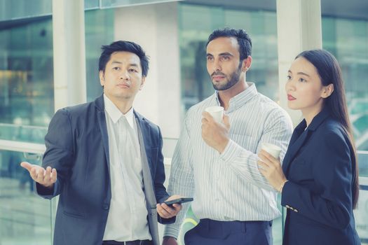 Two handsome young businessmen and lady in classic suits are holding cups of coffee, talking and smiling, standing outside the office building.